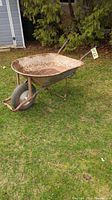 Side and front view of an all metal wheelbarrow on grass showing rusted basin and solid rubber wheel.