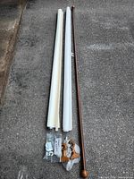 Two rolled white fabric roller blinds and a brown wooden curtain rod laid side by side on ground with mounting hardware placed below blinds and rod.