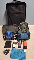 Photo shows the large black rolling suitcase, olive green backpack, black soft briefcase, two blue Coleman cooler bags, three travel cups/thermos, a small black cloth and a blue waterproof bag, along with a black and blue headlamp.