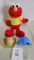 Red plush Elmo with yellow star shirt sitting with tan cowboy hat and blue captain hat in front. Includes advertising card.
