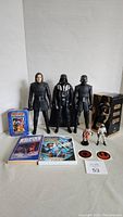 Three large black Star Wars action figures including Darth Vader and two others, two smaller Star Wars figures, six collector cards on left, two stickers in front, three books and Wii case in front