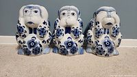 Front view of the three ceramic Wise Monkeys in blue and white floral design arranged side by side on a beige carpet against a gray wall.