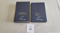 Two navy blue hardcover Ernest Hemingway books side by side on a light surface, showing front covers with gold and silver embossed titles and signature.