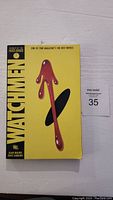 Front cover of Watchmen graphic novel showing yellow cover with iconic blood splatter design and black spine with title text.