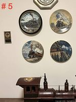Wall-mounted photo showing four round collectible locomotive plates with steam engine themes, each about 9.5 inches in diameter, good condition, along with a small vintage object and a model train component beneath.