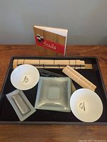 Full sushi set contents arranged in black tray showing bowls, plate, rolling mats, chopsticks, sushi knife, and recipe book.