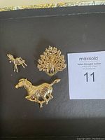 Two gold-tone horse brooches and a gold-tone peacock brooch on black surface with auction number.