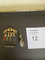 Eleven assorted costume jewelry brooches on a dark surface, including a gold tone MOMO pin with colorful child figures and other varied designs.