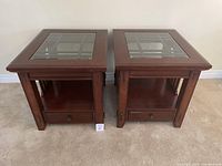 Pair of two wooden end tables with glass inset tops, shown side-by-side on carpeted floor.