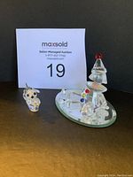 Two Swarovski crystal figurines, a puppy and a clown on an oval mirror base, positioned with white maxsold auction card behind them.