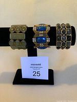 Three costume jewelry bracelets displayed on a black stand including a green-toned bracelet with rounded stones, a gold-tone bracelet with blue stones and rhinestones, and a greyish stone linked bracelet.
