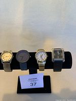 Four watches displayed on black holder with varying strap materials and dial styles.