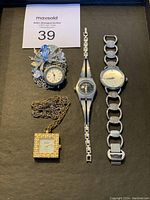 Top view of entire lot showing the brooch pin with blue floral design and embedded clock, two silver-tone bracelet watches with linked bands, and a gold-tone square pendant necklace watch with rhinestone border.