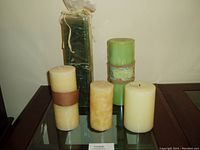 Five assorted large candles arranged on glass table, showing color and size variation including a wrapped rectangular green candle and one used candle