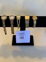 Photo of five assorted metal bracelet watches displayed on a black watch stand, showing varying shapes and band styles.