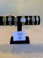 Full view of all 7 assorted metal bracelet watches displayed on black stand with maxsold lot number card.