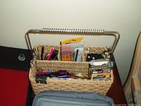 Woven desk caddy with assorted pens, pencils, staples, and other small office supplies. The caddy has a metal handle and multiple compartments.