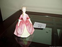 Front side view of Royal Doulton porcelain figurine of a woman in a pink and white dress on a glass table.