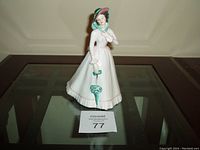 Front view of Royal Doulton porcelain figurine Julia holding a green parasol and a hat, standing on a glass table.