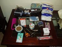 Photo shows assortment of toiletry and personal care items laid out on table, including pink comb, cotton balls, scissors, curlers, hair dryer, and various small boxes and packets.
