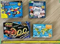 Photo showing four boxed children's toys arranged on a wooden floor, including Aero-trax Plane, Rapid Doodle magnetic board, Speed Racer car track, and a world map jigsaw puzzle.