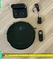 Top view of Eufy RoboVac, charging dock, power adapter, and remote control with measuring tape showing approximately 12 inches diameter of vacuum