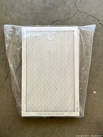 Single Filtrete furnace filter wrapped in clear plastic packaging lying flat on concrete floor, showing white filter and frame.