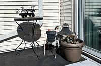 Black metal round table with a black metal decorative plant hanger on top, white metal wire plant stand, large brown plastic planter, black rustic lantern, and terracotta pot on balcony floor.