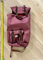 Front view of burgundy Herschel backpack with brown leather straps and front pocket. Measuring tape alongside shows approximate height of 25 inches.