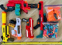 Photo shows six Nerf blasters and two bags of darts and components arranged on wooden floor