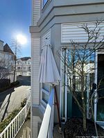 Closed beige patio umbrella attached to brown pole leaning against outdoor railing next to a building and glass door.
