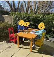Three children seated outdoors at the solid wood rectangular craft table with four colorful wooden chairs in red, yellow, blue, and green.