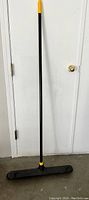 Image of the heavy duty XL push broom against a white door and wall, showing full length, black handle with yellow accents, and wide black bristle broom head.