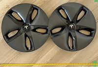 Photo showing two Tesla Model 3 18-inch aero wheel covers laid on wood flooring, displaying dark gray aerodynamic design with glossy black insets and Tesla logo center caps. Yellow tape measure below shows 18 inches length for scale.