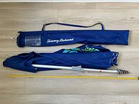 Tommy Bahama navy blue beach umbrella folded with aluminum pole and spiral sand stake. Navy blue carrying case with shoulder strap and logo included. Yellow measuring tape alongside shows approximate length of umbrella and case.