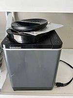 Front and side view of the compact food recycler showing the gray body, black top lid with vent holes and paper sheet under lid.
