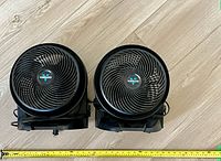 Front view of two black Vornado air circulator fans side by side on wood floor, power cords visible, with tape measures showing approximately 16 inch diameter each.