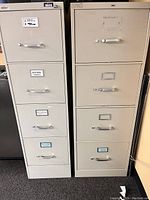 Two tall metal filing cabinets, each with four drawers with handles and label holders. One cabinet is slightly lighter colored than the other.