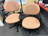Two tan fabric office chairs with black armrests and black rolling bases on carpet floor near desk.