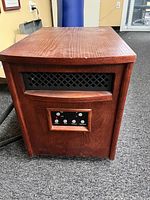 Front view of a compact electric space heater with a wooden exterior. Shows a black vent grill on the upper section and digital control buttons on a small display on the lower section of the front panel.