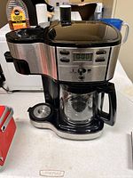 Front view of Hamilton Beach coffee maker showing stainless steel and black plastic design, glass carafe with handle, digital clock, and buttons.