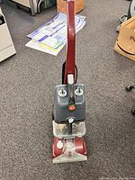 Front view of a red Hoover electric carpet cleaner on carpet, showing the handle, dual tanks, and transparent cleaning head.