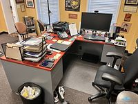 Photo of large L-shaped office desk with reddish-brown wood finish top and grey base, cluttered with office items (not included in sale). Shows left and angled corner section.