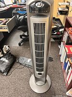 Full front-facing view of an oscillating tower fan showing vertical vents, control panel at the top, and circular base. The fan appears to be in an office or storage room area.
