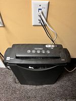 Black Mainstays paper shredder plugged into a wall outlet showing the top control panel and paper slot for shredding.