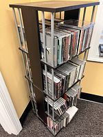 Photo shows a tall rotating CD rack with metal sides and wooden shelves filled with CDs, demonstrating its capacity and design.