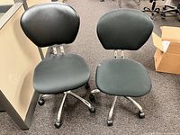 Two black rolling office chairs side by side on office carpet, next to desk and cardboard box.