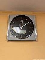 Front view of square wall clock with silver frame, black clock face, and digital display at bottom for day, date, and temperature.