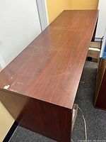 Photo showing top and side view of a dark brown wooden office table, approximately 6 feet long, placed against a wall with minor surface wear visible.