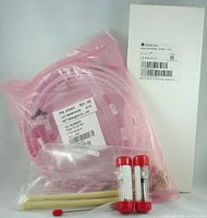 Clear flexible tubing with labels, two red-capped cylindrical components, small plastic bag with components, and white labeled box displayed together